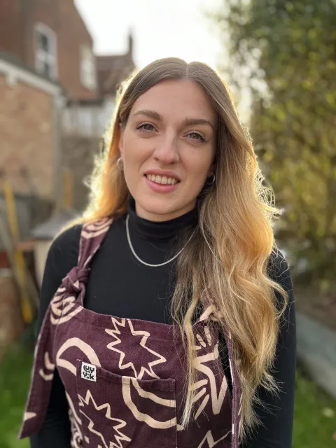 A woman with long blond hair smiling standing outside with tree and building behind her